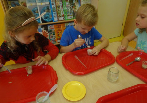 Troje dzieci siedzi przy stoliku, obserwują reakcje wody utlenionej z drożdżami w swoich pojemnikach.
